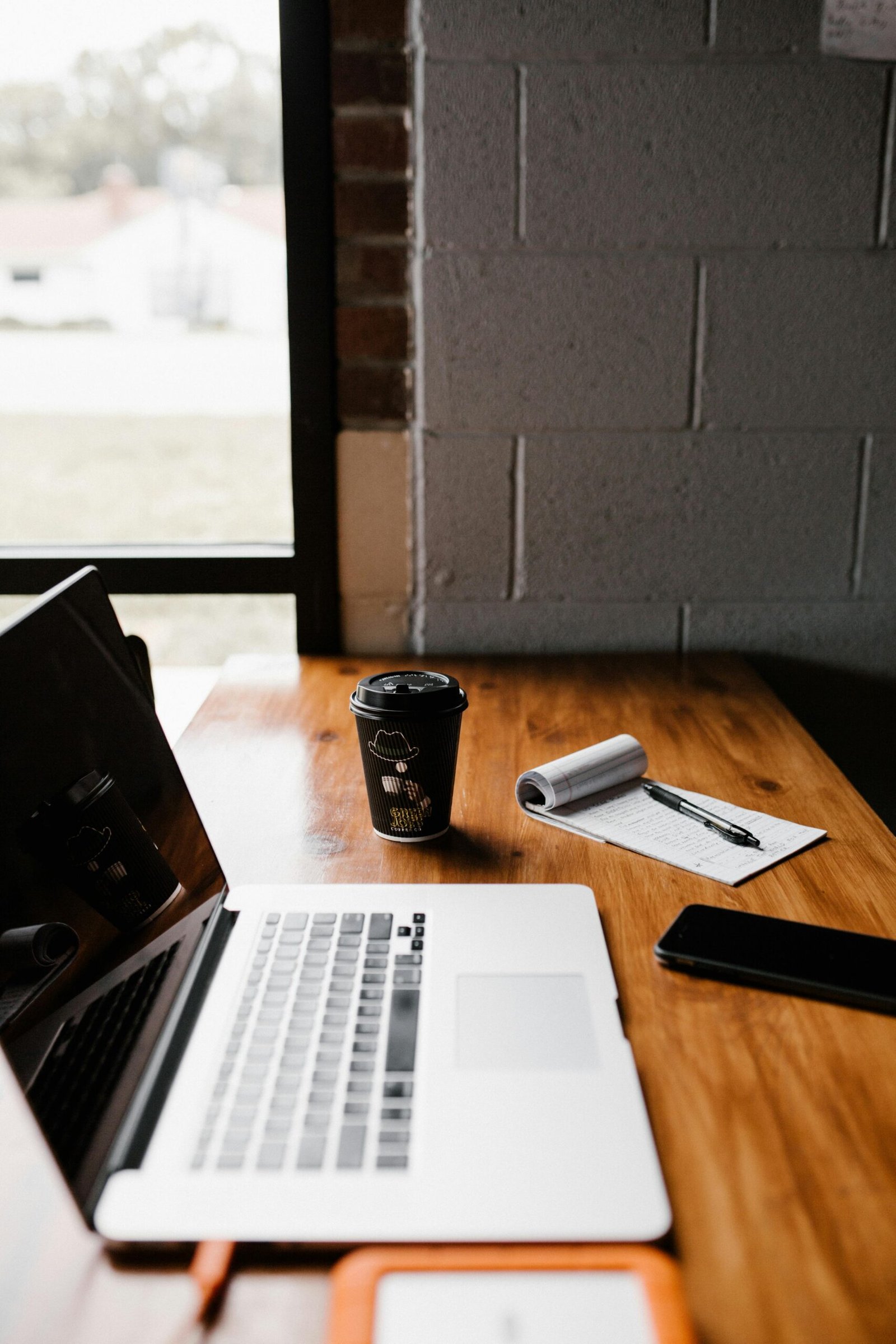 Modern office desk setup with laptop, coffee cup, and notepad by window.